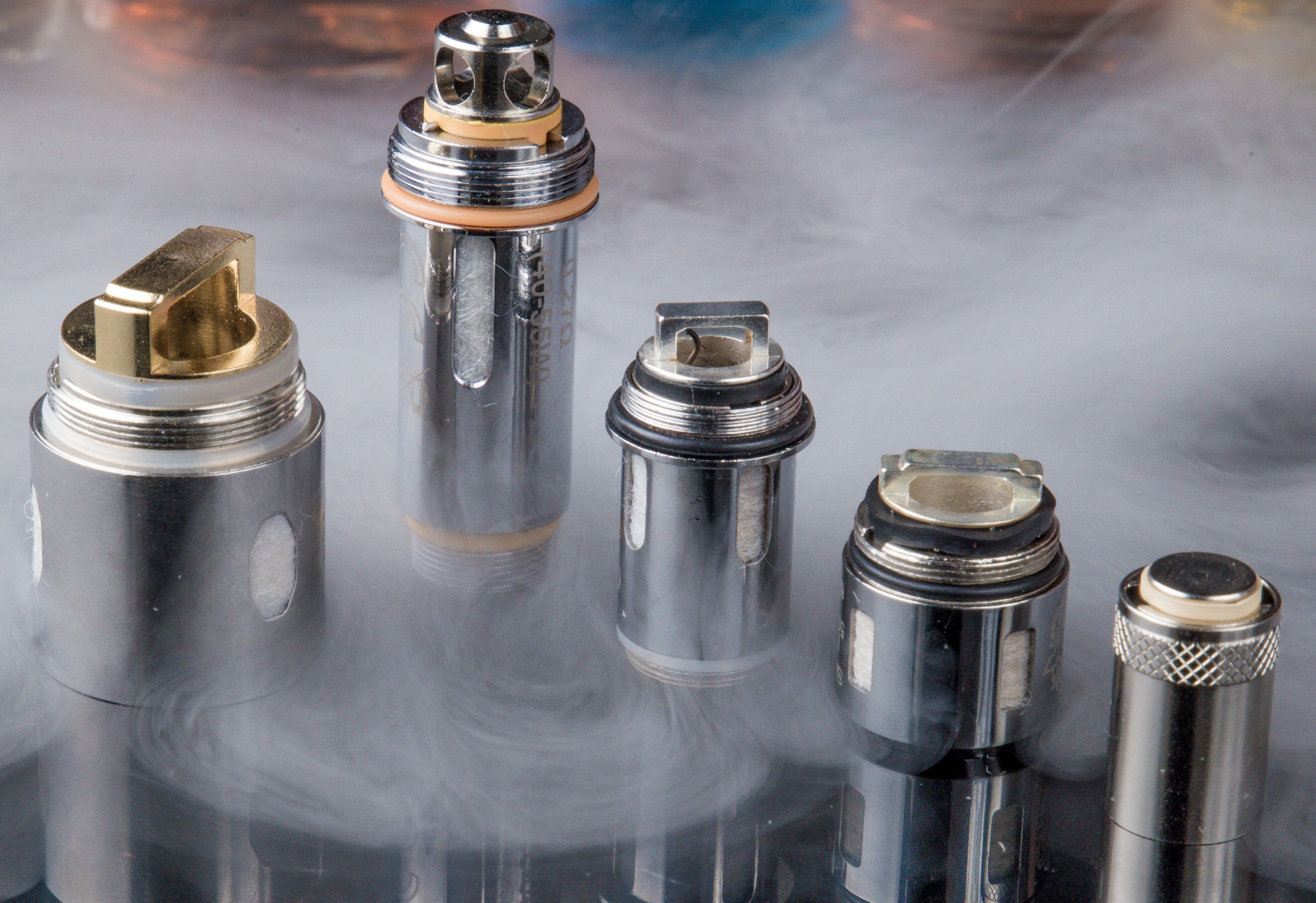 Five different silver vape coils standing in a cloud of vapor on a reflective surface.